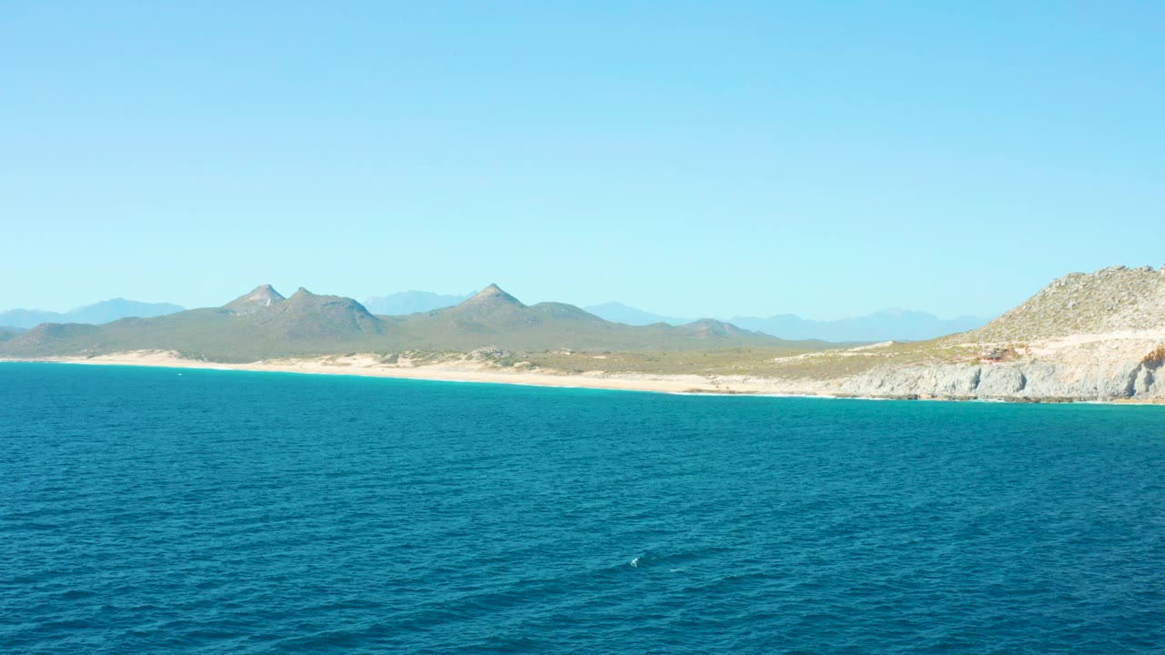 A wide East Cape coastal frame shows turquoise sea, pale beach, rugged distant hills, and the rocky right edge of the shore, with no visible people or structures.