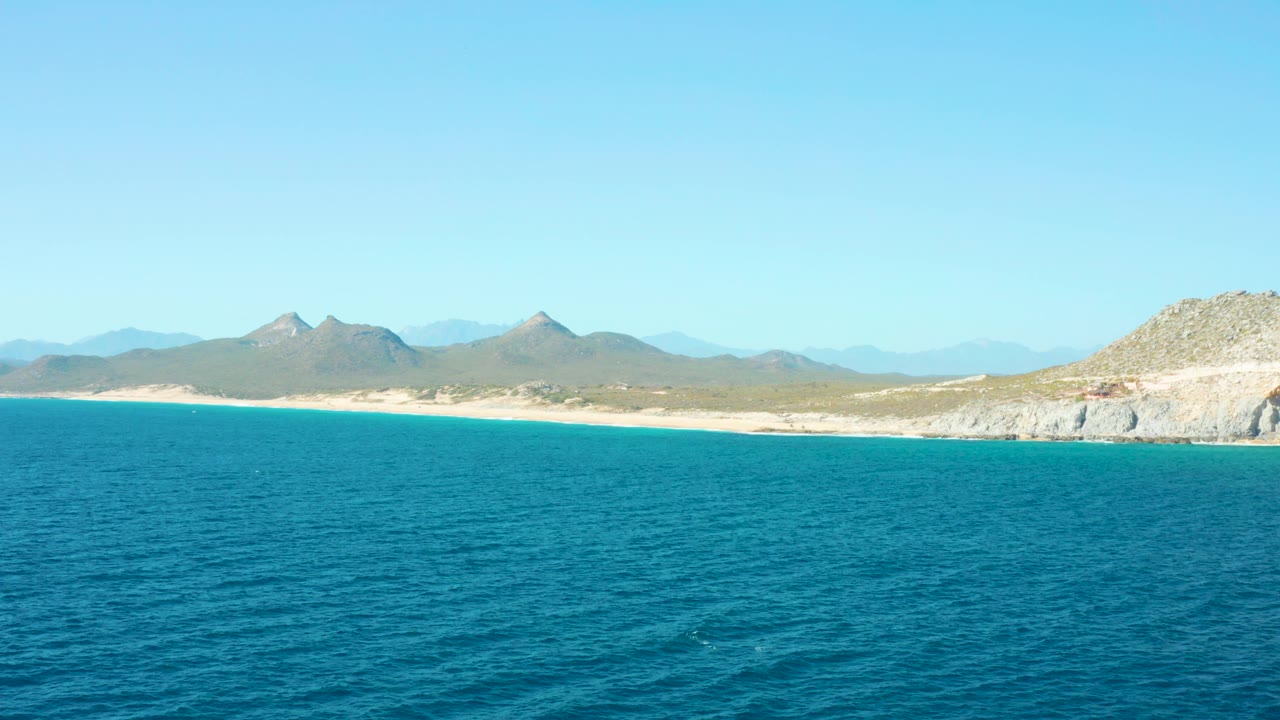 East Cape blue water stretches toward a sandy beach and mountain backdrop, with the rocky point more prominent at the right edge and no visible people.