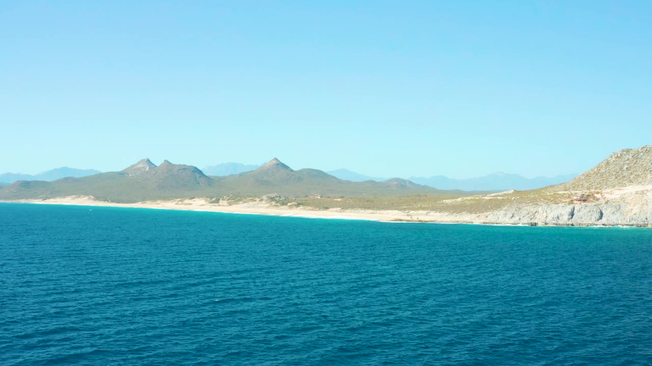 A broad East Cape BCS Mexico shoreline stretches behind rippled blue water, with pale sand, dry mountain ridges, a rocky right edge, and no visible people.