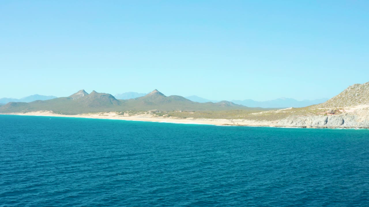 Blue ocean occupies the lower frame while the East Cape BCS Mexico beach and mountain line remain distant under bright clear sky, with no visible people or boats.