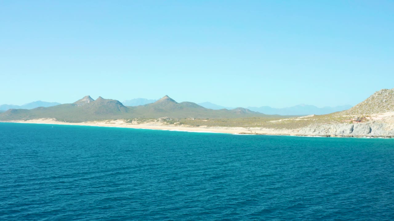 The East Cape BCS Mexico coast appears across textured blue water, with a low sandy beach, hazy mountain backdrop, rocky right shoreline, and no visible people.