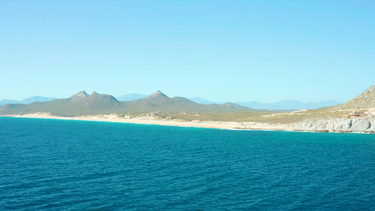 Wide ocean, sandy coast, and distant mountains under clear sky.