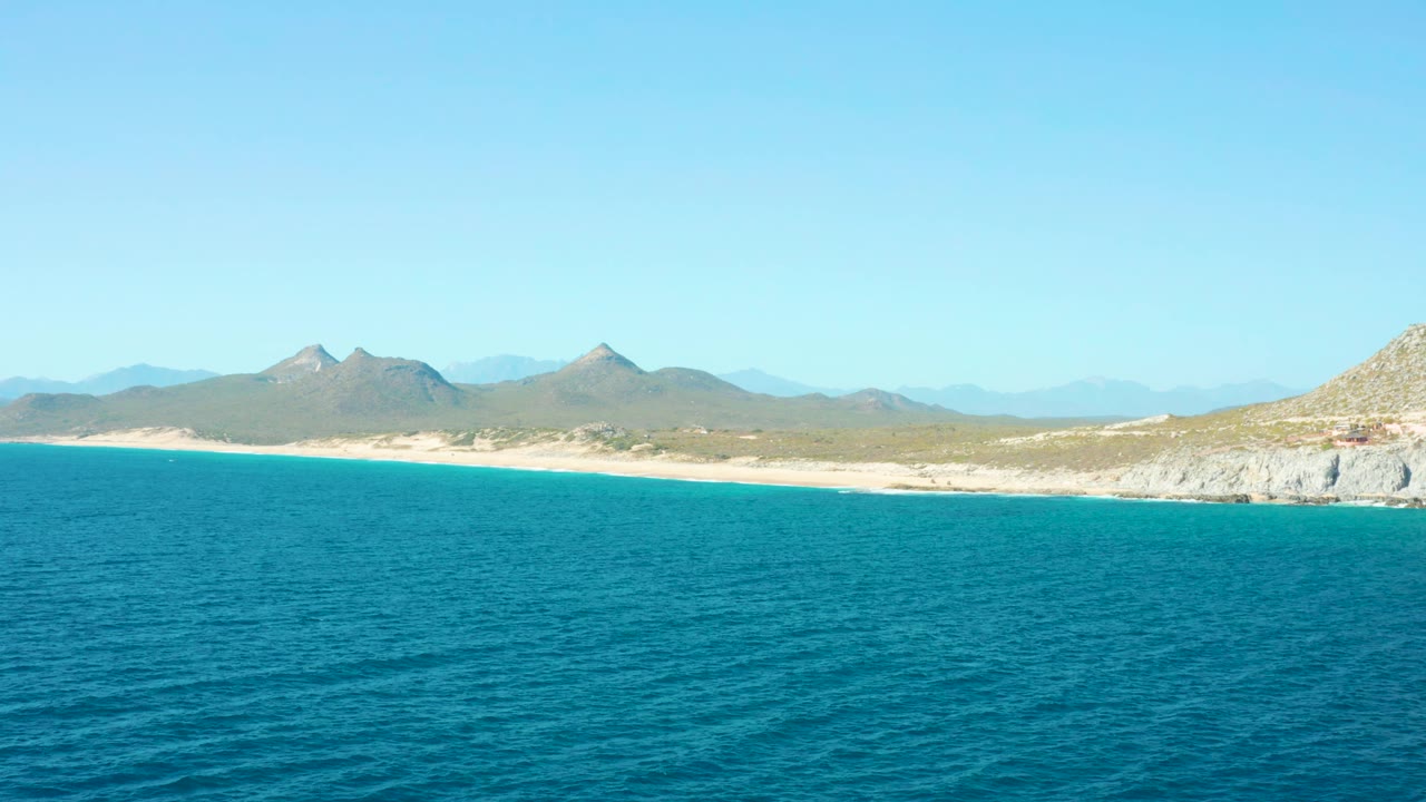 At 0:01, the blue water surface sits below a long pale beach and mountain backdrop in East Cape BCS Mexico, with no visible people.