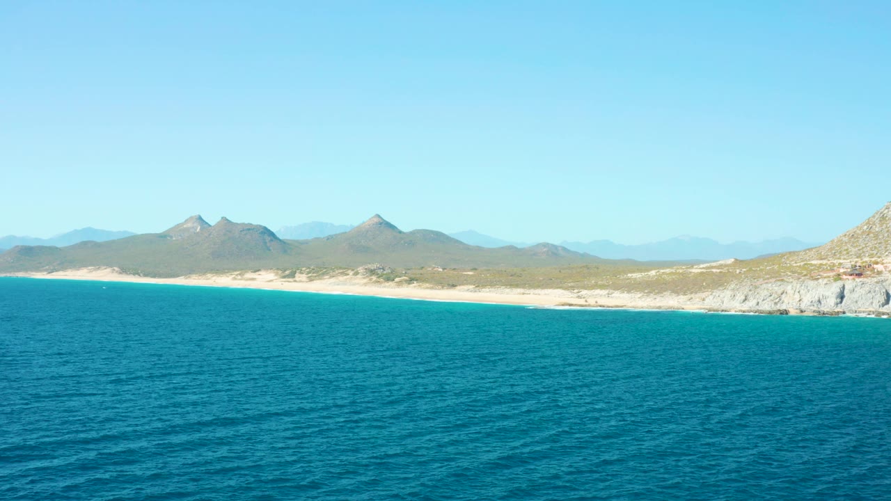At 0:02, rippled turquoise ocean leads to a pale East Cape BCS Mexico beach, with a rocky point at the right edge and mountains behind, with no visible people.