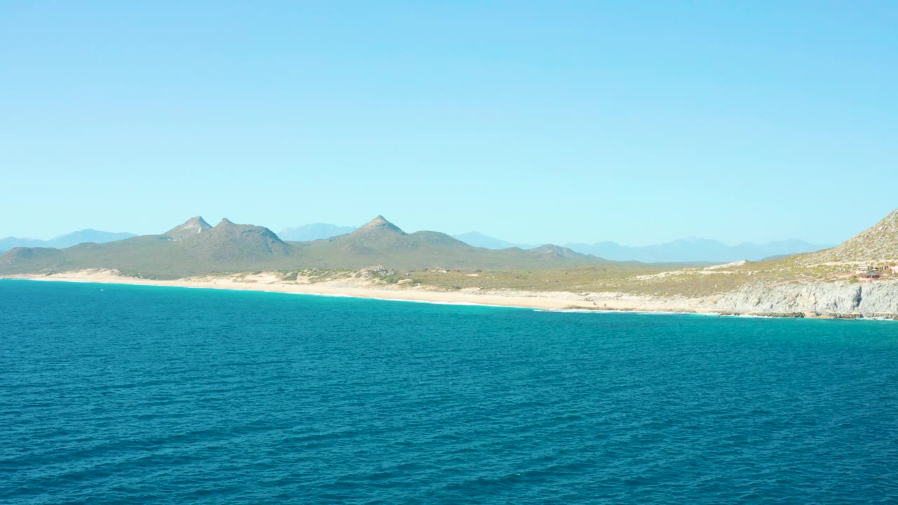 At 0:03, the East Cape BCS Mexico coast appears as a narrow sand band between calm blue water and hazy inland mountains, with no visible people.