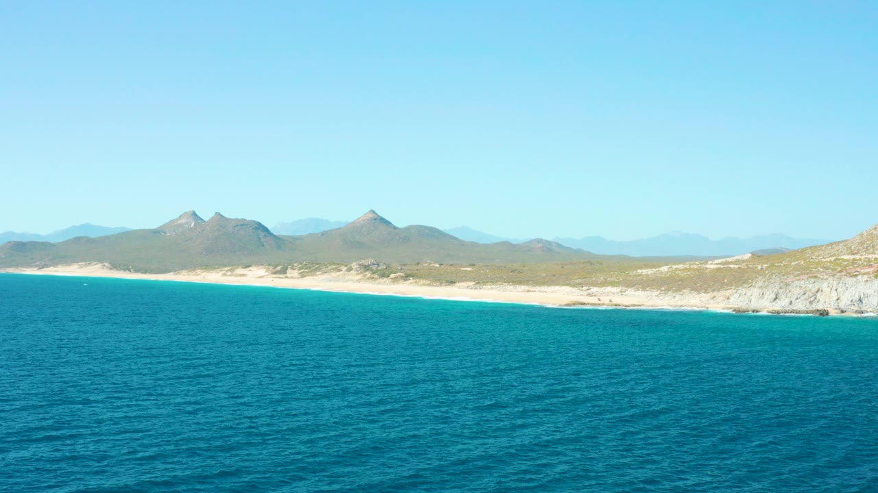 Wide aerial view of East Cape BCS Mexico with turquoise water in the foreground, a sandy shoreline, hazy mountains, and no visible people.