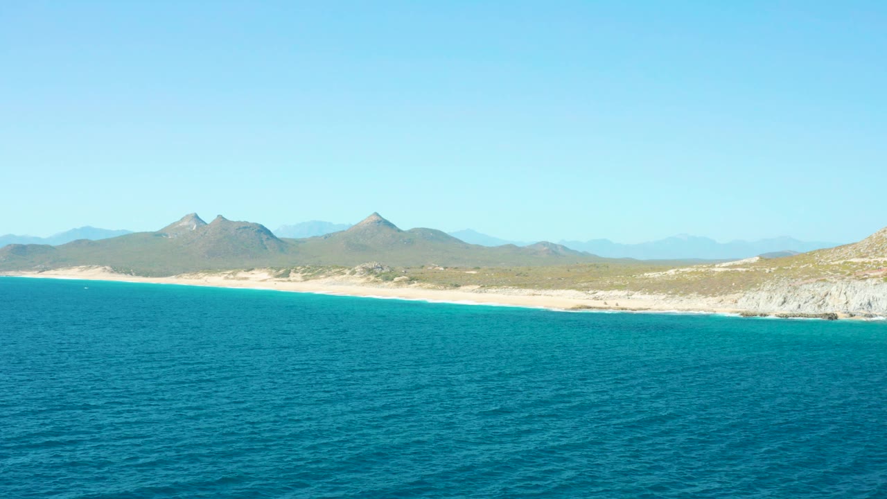 Aerial coastal frame from East Cape BCS Mexico showing textured blue water, a thin sand strip, pale coastal rock, distant mountains, and no visible people.
