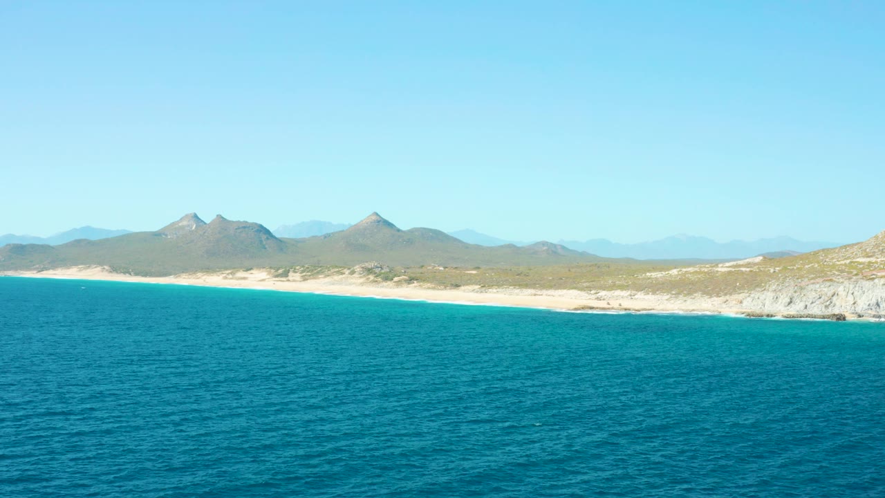 East Cape BCS Mexico shoreline with calm turquoise sea, light sand, low coastal scrub, a pale bluff at right, hazy mountains, and no visible people.
