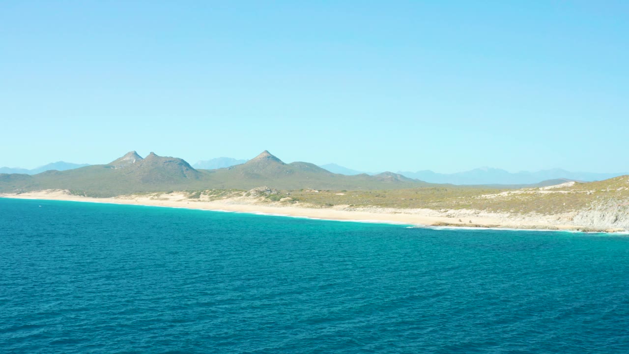 Mid-clip frame showing blue ocean in the foreground, a pale beach strip, sparse coastal scrub, distant peaks, and clear sky over East Cape, with no visible people.