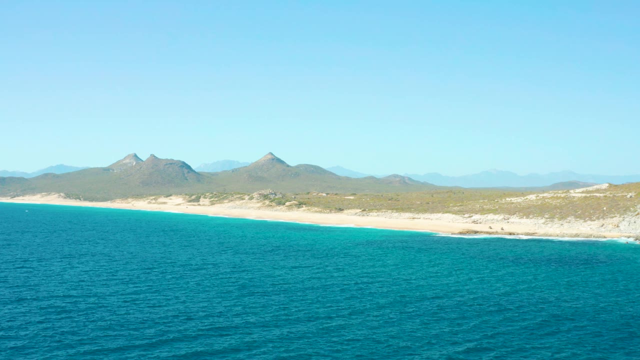 Wide aerial frame over East Cape BCS Mexico showing rippled turquoise water, a pale sandy beach, dry coastal vegetation, distant mountain peaks, clear sky, and no visible people.