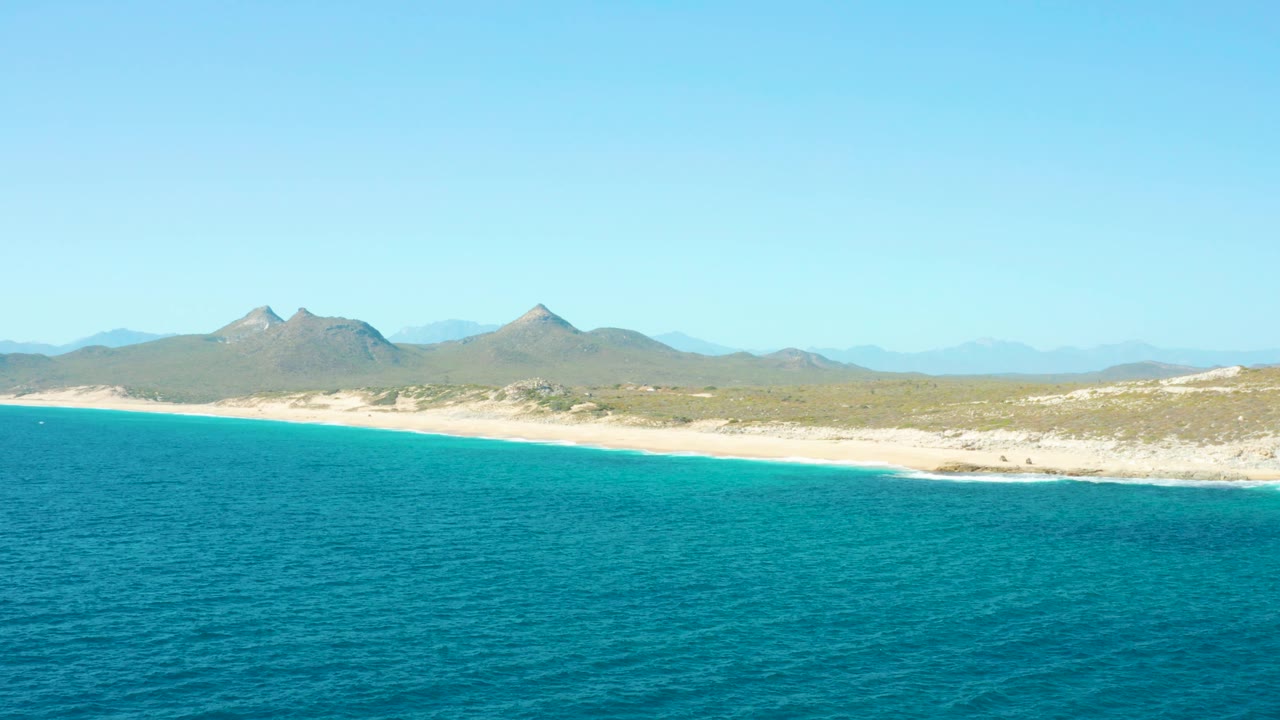 East Cape BCS Mexico aerial frame with textured turquoise sea in the foreground, beach and scrubland across the middle, a mountain ridge in the background, clear sky, and no visible people.