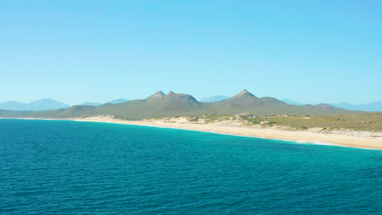 Wide aerial view of East Cape BCS Mexico with turquoise ocean in the foreground, pale beach along the shore, distant mountains under clear sky, and no visible people.