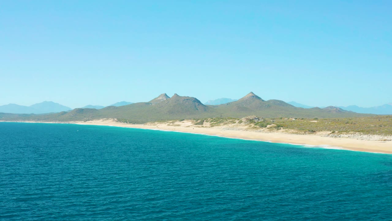 East Cape BCS Mexico coastline seen from above, with blue-green water filling the foreground, a narrow sandy shore, low inland scrub, mountain peaks, and no visible people.