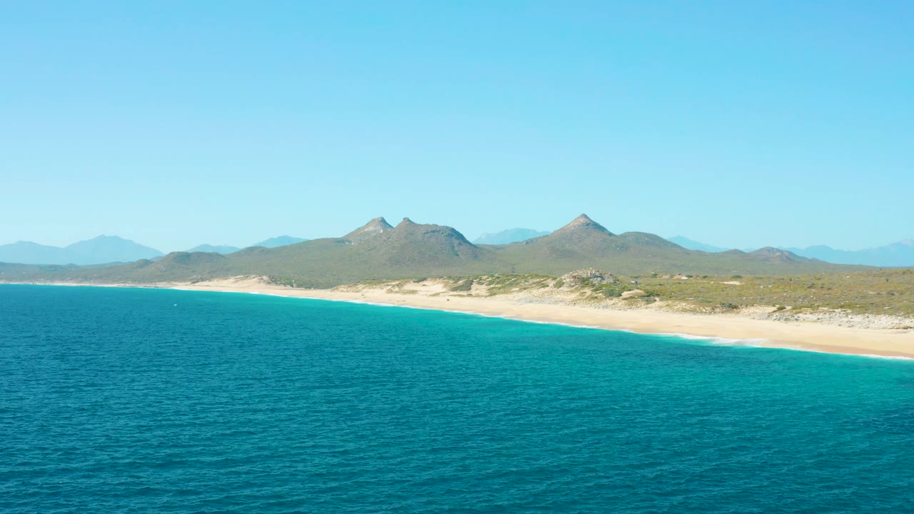 Aerial East Cape BCS Mexico beach view with textured turquoise ocean in front, a bright strip of sand at midframe, inland brush, distant mountains, and no visible people.
