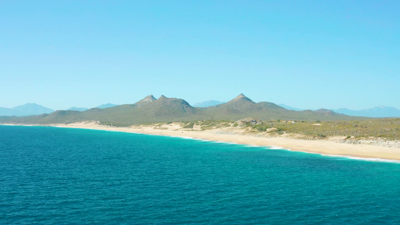 Aerial view of East Cape BCS Mexico with turquoise ocean in the foreground, a pale sandy shoreline, low desert vegetation, distant mountains, clear blue sky, and no visible people.