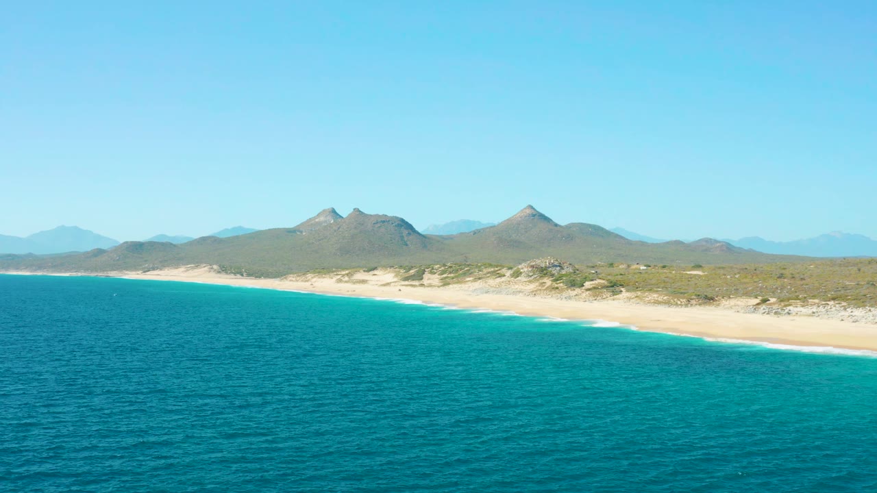 Wide aerial frame over East Cape BCS Mexico showing rippled turquoise water, a thin line of white surf, sandy beach, scrubby inland terrain, distant mountains, and no visible people.