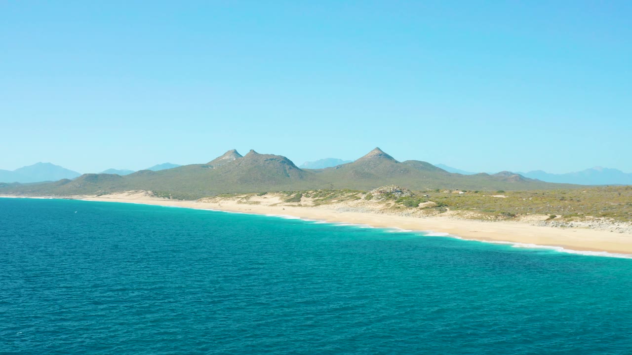 Aerial shoreline view in East Cape BCS Mexico with teal ocean water closest to camera, light sand along the coast, low brush inland, layered mountains in the background, and no visible people.