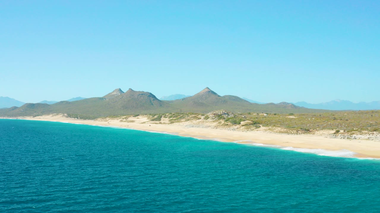 Wide aerial view of East Cape BCS Mexico with turquoise ocean in the foreground, pale sand along the shore, distant mountains under clear sky, and no visible people.