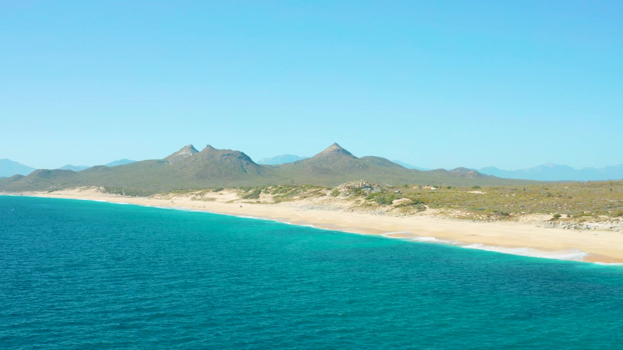 A wide East Cape BCS Mexico beach view shows turquoise sea in front, an empty sandy shoreline, low dune vegetation, distant mountain peaks, and no visible people.