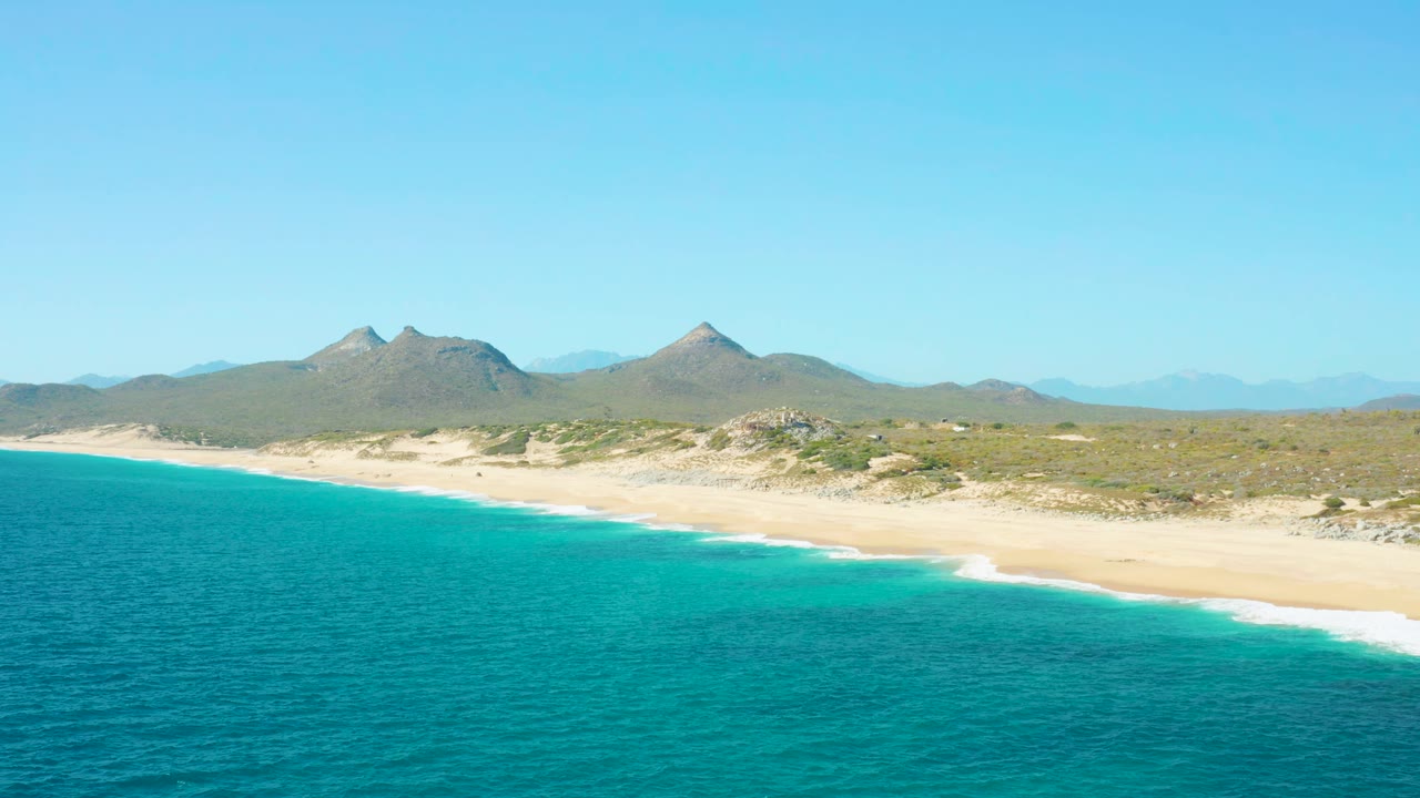 Aerial frame over East Cape BCS Mexico showing a longer band of turquoise water against pale sand, scrub-covered land behind it, two pointed mountain forms, and no visible people.