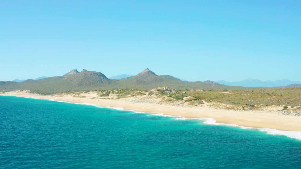 Aerial frame from East Cape BCS Mexico with turquoise sea occupying the lower half, a thin white surf edge, sandy beach, inland scrub, distant hills, and no visible people.