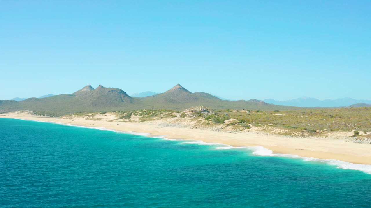 Mid-clip aerial frame over East Cape BCS Mexico with the shoreline angled across the scene, turquoise water below, scrubby dunes and low mountains beyond, and no visible people.
