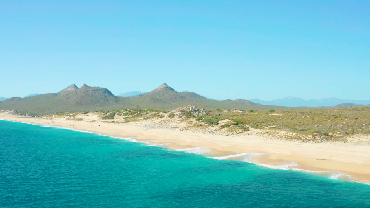 Aerial view over East Cape, Baja California Sur with teal ocean taking more of the lower frame, thin white surf lines on the sand, scrubby dunes, distant mountains, blue sky, and no visible people.