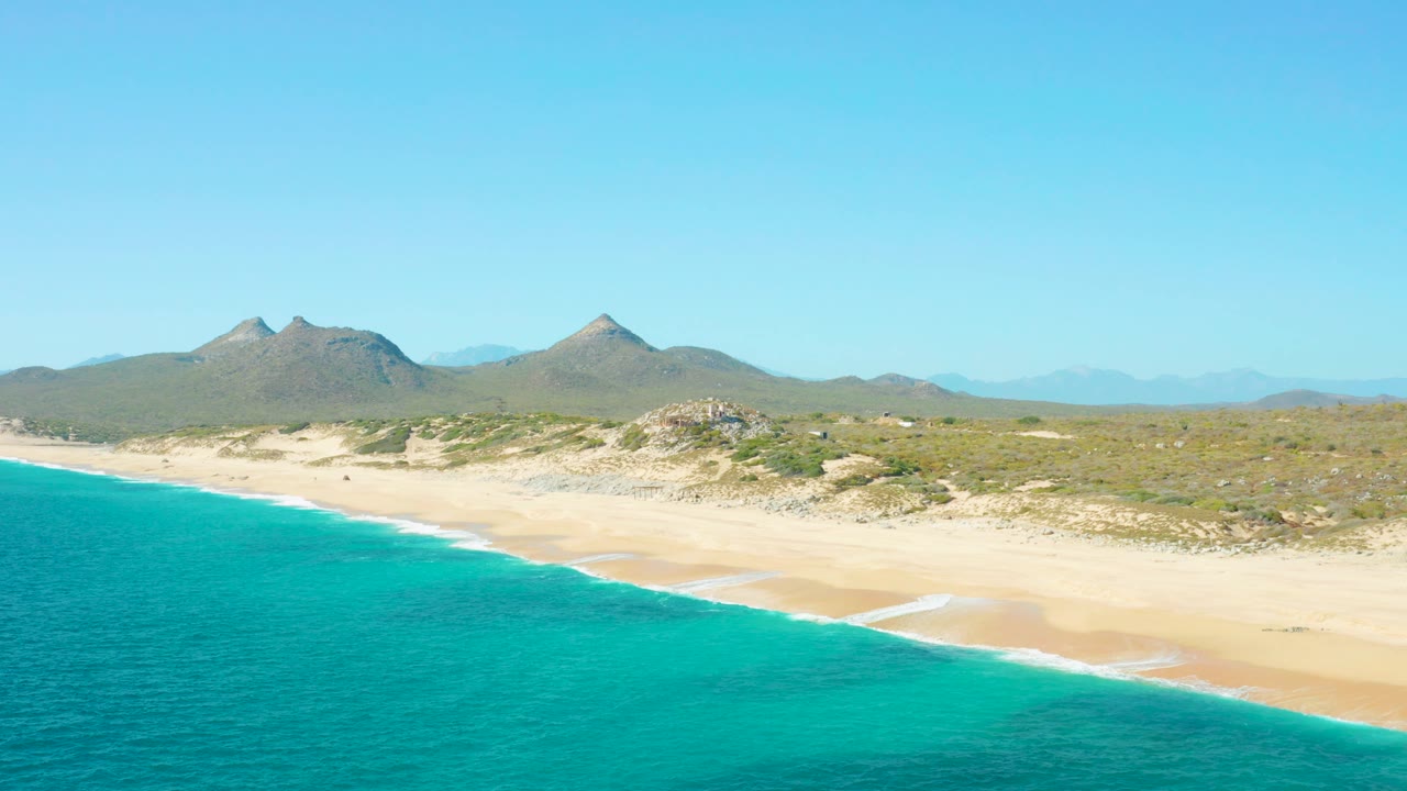 Mid-clip aerial frame from East Cape, Baja California Sur showing blue-green water at left, a narrow white wave line on the sand, dry dune scrub behind the beach, hazy mountains inland, clear sky, and no visible people.