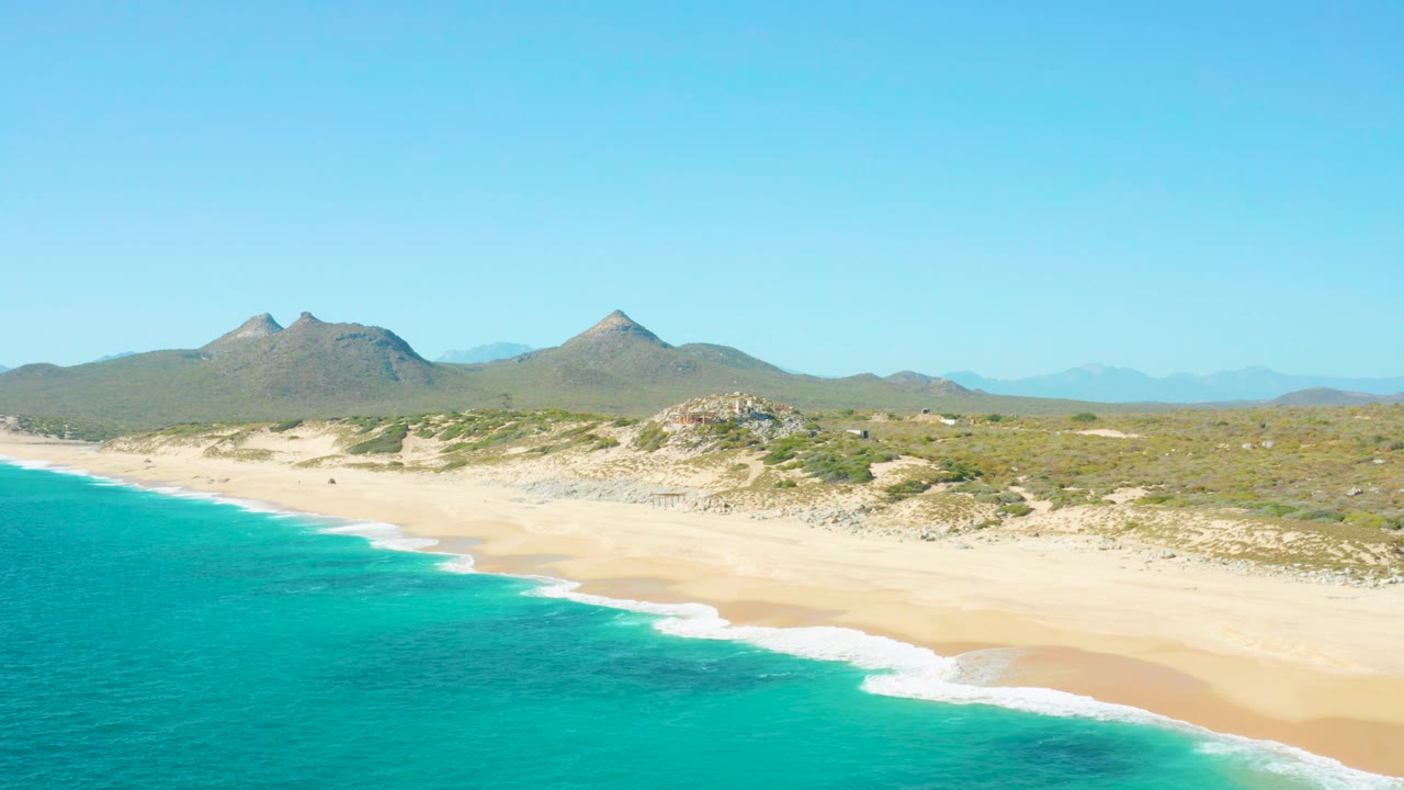 Wide sandy shore with turquoise waves and mountains under clear sky.