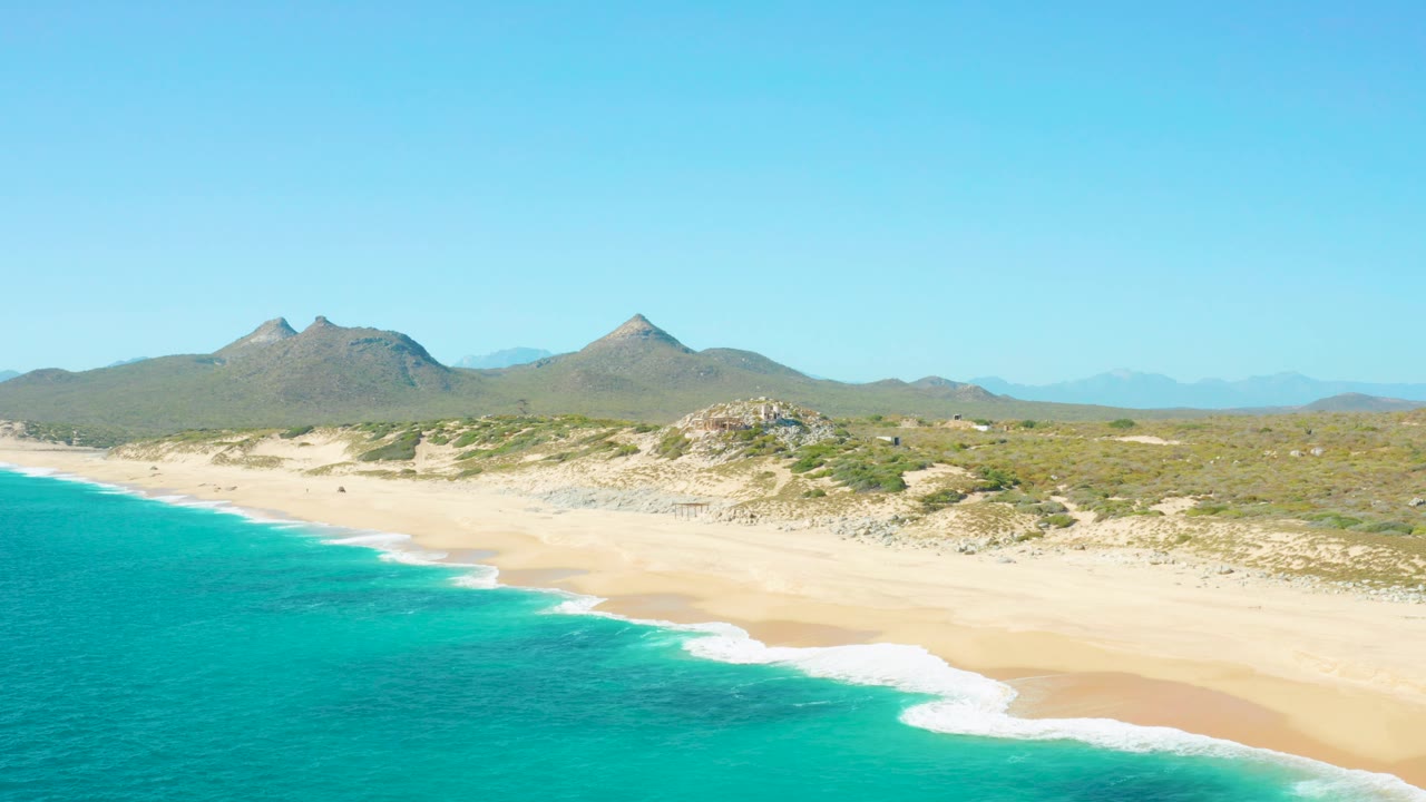 Aerial frame over East Cape showing brighter turquoise water, a clean strip of white surf, broad beige sand, low coastal vegetation, distant mountains, and no visible people in this frame.