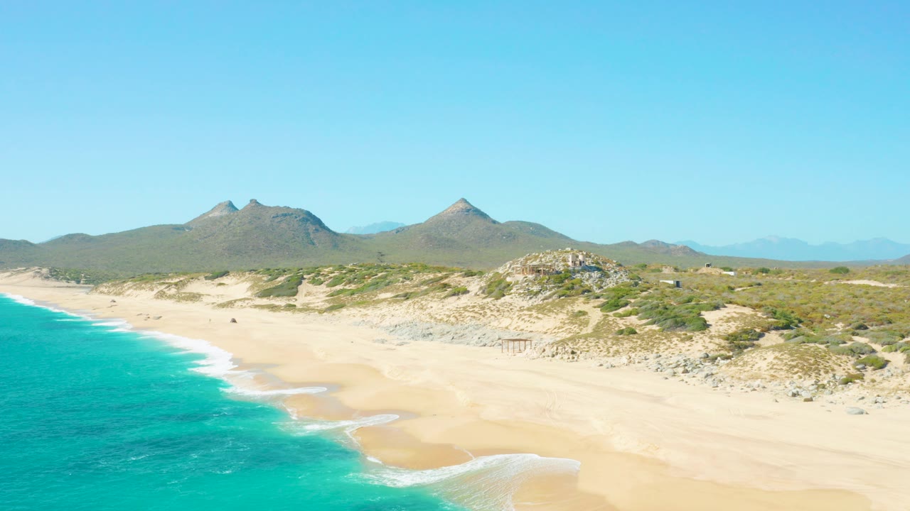 A later aerial frame shows more of the curved surf line on the East Cape BCS Mexico beach, with turquoise water in the lower left, dunes to the right, and no visible people.