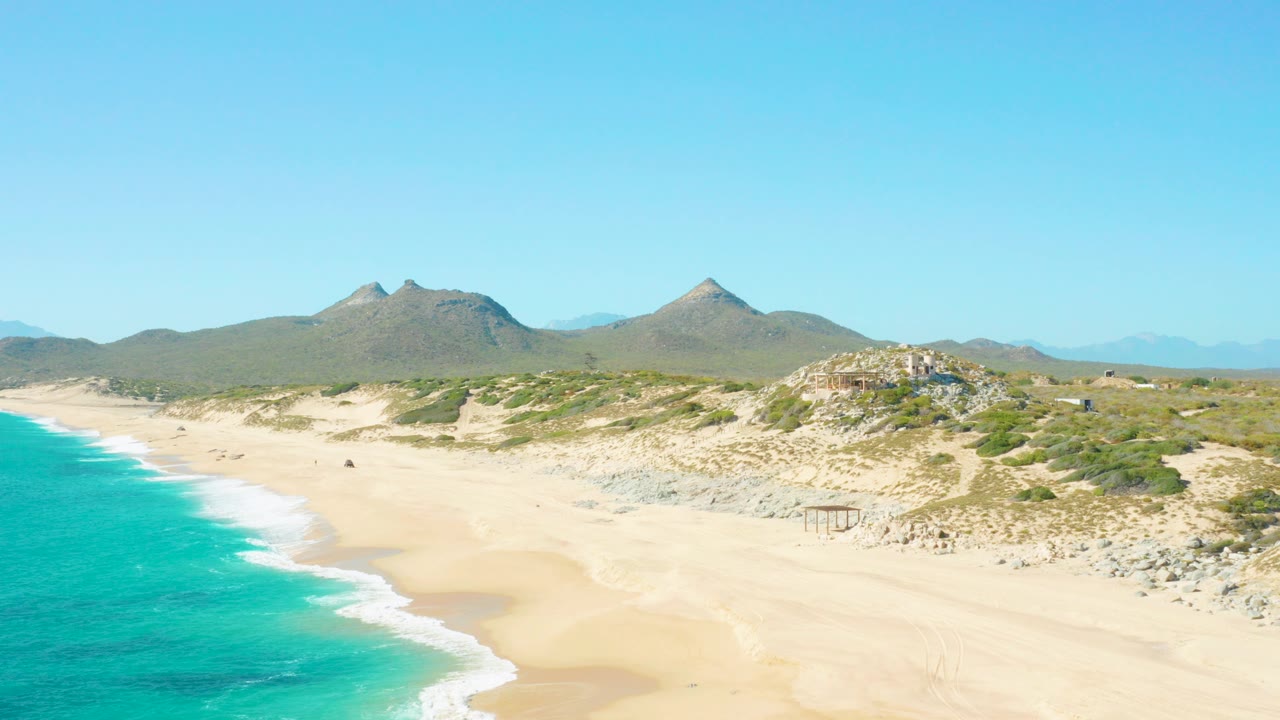 Wide aerial view of an East Cape, Baja California Sur beach with turquoise water at left, pale sand and dunes in the center, low mountains beyond, and no visible people in this frame.