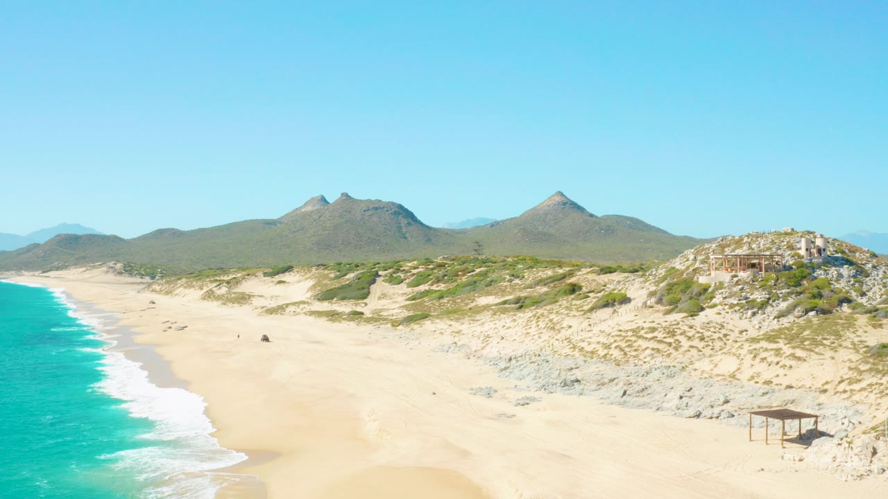 Wide aerial view of an East Cape BCS Mexico beach with turquoise surf on the left, pale sand across the center, low dunes, rocky terrain, distant mountains, and tiny beach figures visible.