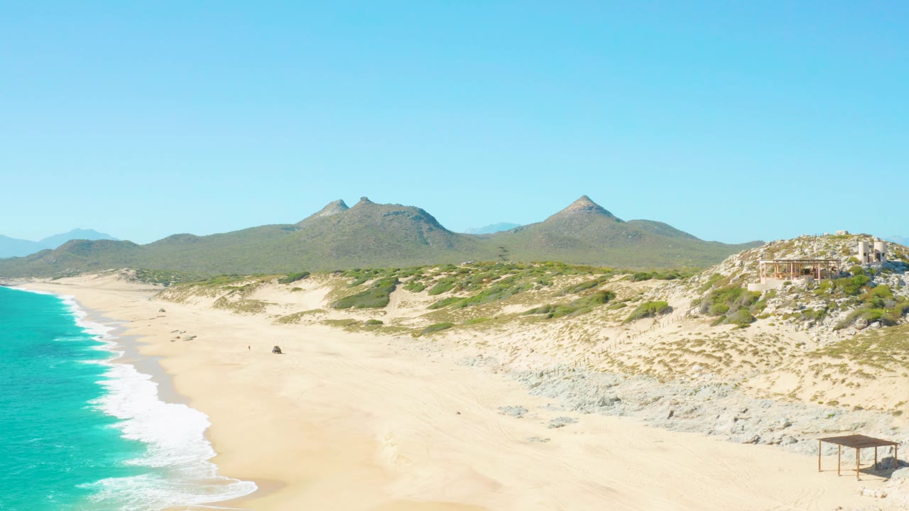 Wide East Cape BCS Mexico coastal frame with the surf shifted farther left, sand and dunes filling most of the view, rocky hills at right, distant mountains, clear sky, and small beachgoers visible.