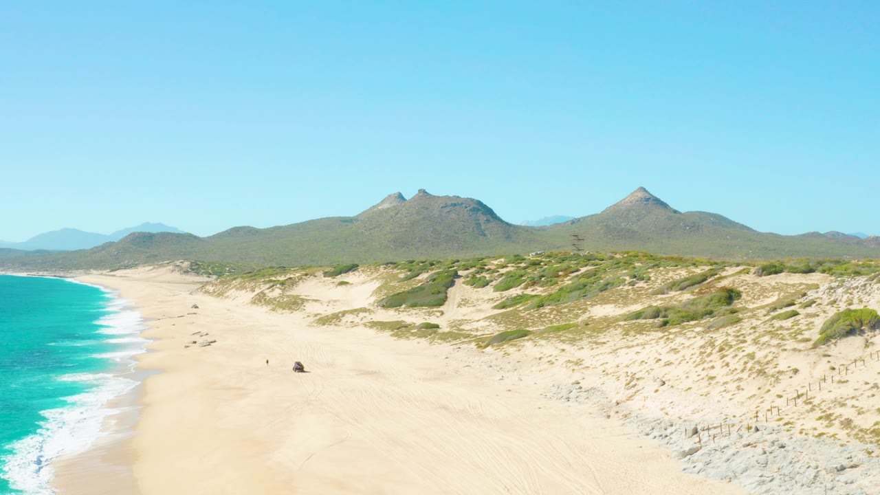Aerial view in East Cape BCS Mexico showing turquoise surf on the left, a wide sandy beach, dune vegetation, distant mountains, and small people visible on the sand.