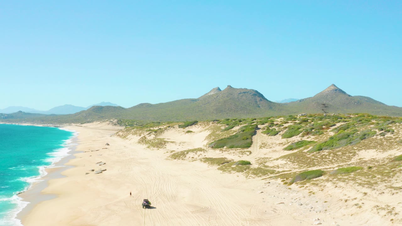 Aerial view of East Cape BCS Mexico with turquoise surf at left, pale sand dunes, green scrub, distant mountains, a small vehicle near the lower beach, and a tiny person on the sand.