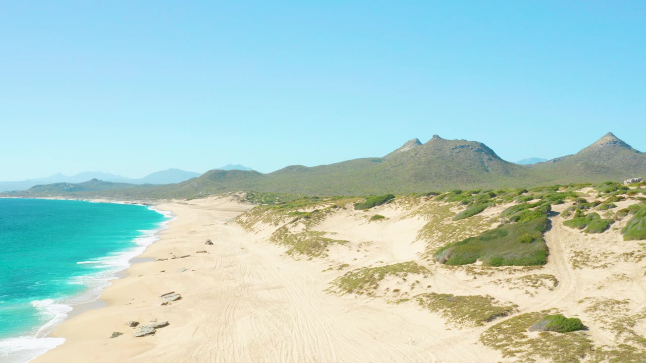 At 0:00, East Cape BCS Mexico shoreline fills the frame with turquoise water on the left, pale sand and dune vegetation in the center, distant mountains under clear sky, and no visible people.