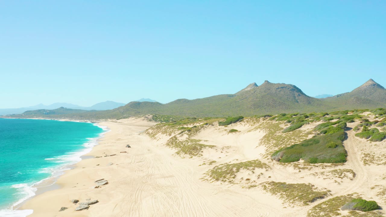 At 0:02, East Cape BCS Mexico dunes and beach dominate the foreground while turquoise water runs along the left edge, mountains sit across the horizon, and no visible people appear.