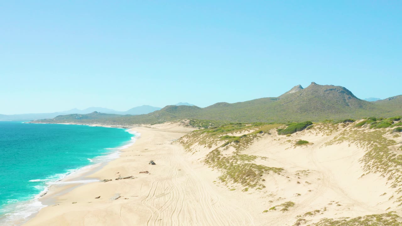 Aerial view over East Cape, Baja California Sur, with turquoise ocean at left, pale sand and dune vegetation at right, distant mountains under clear sky, and no visible people.