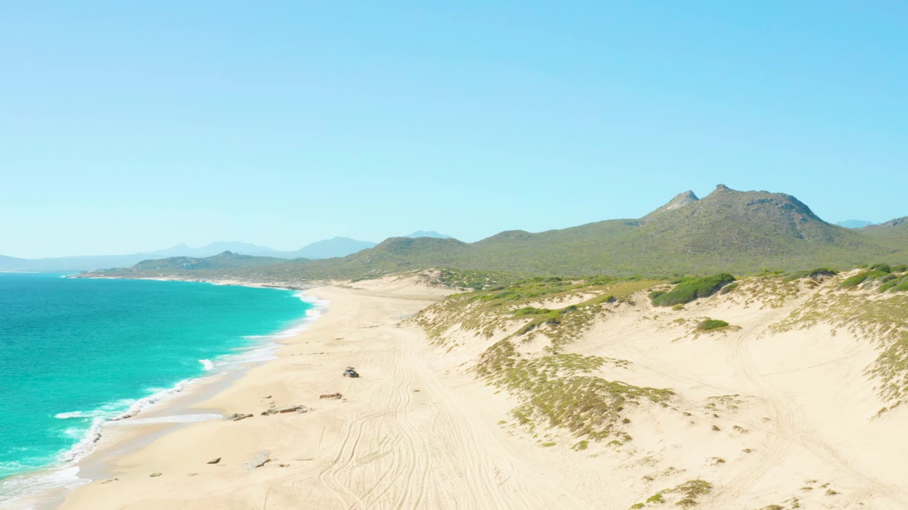 Aerial coastline view in East Cape, Baja California Sur, with turquoise surf on the left, broad pale sand through the center, dune vegetation on the right, distant mountains, and no visible people.