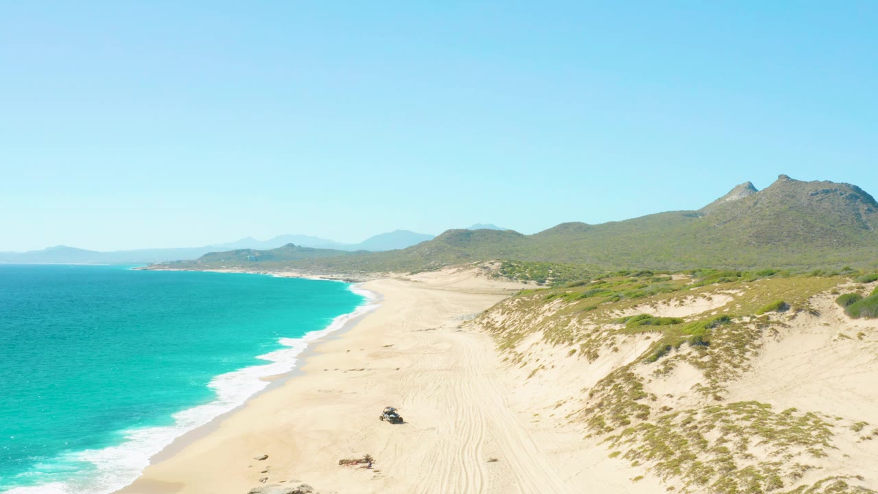East Cape, Baja California Sur beach aerial showing a curving turquoise shoreline, pale sand tracks, dune vegetation, mountains in the distance, a dark vehicle near the lower beach, and no visible people.
