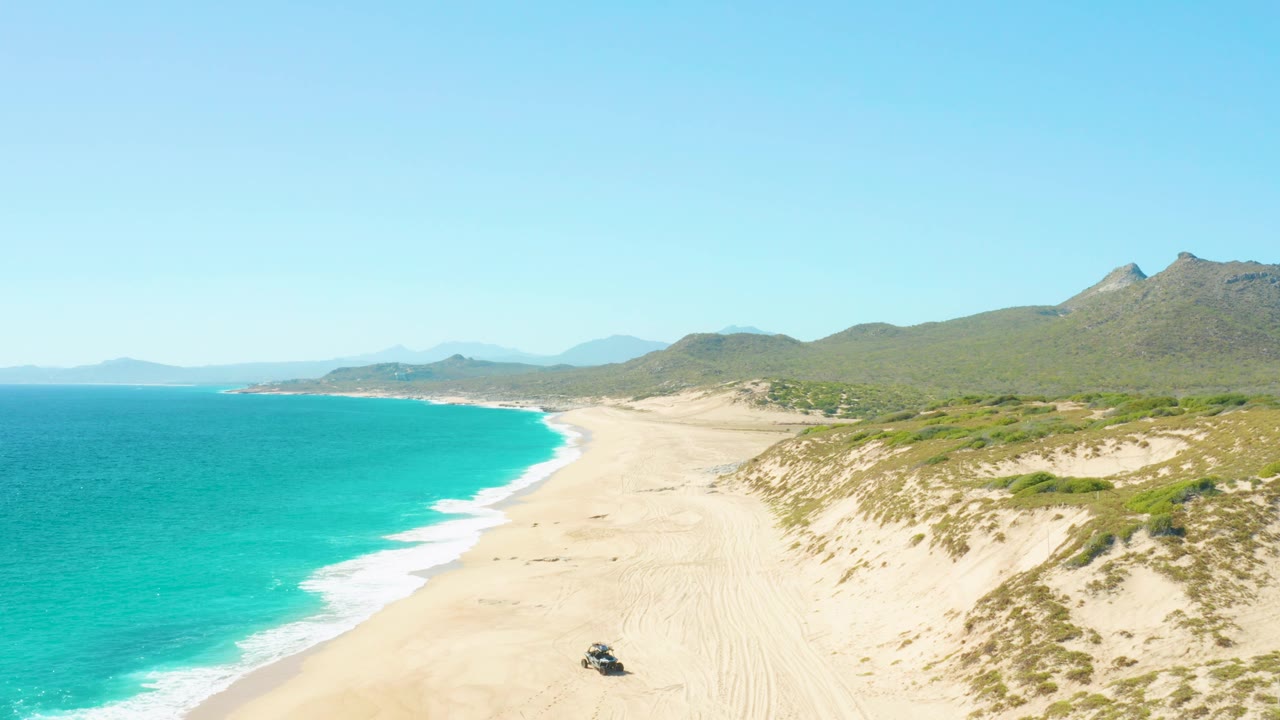 East Cape, Baja California Sur aerial view with the turquoise sea at left, bright surf line, broad sandy beach, scrub dunes, mountains behind the shore, a vehicle near the lower edge, and no visible people.