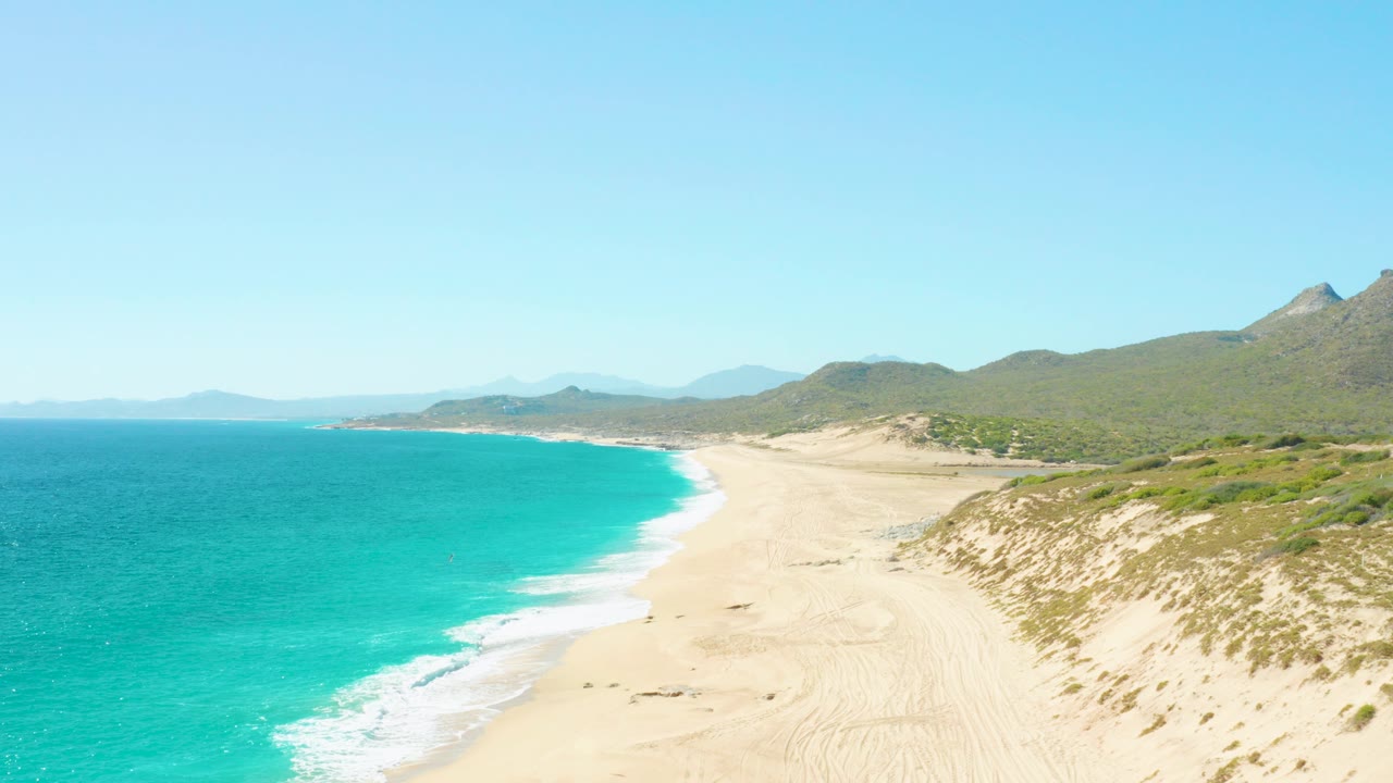 Wide aerial frame over East Cape BCS Mexico showing turquoise ocean on the left, a pale sandy beach, dune vegetation, green hills, distant landforms, and small beachgoers on the sand.