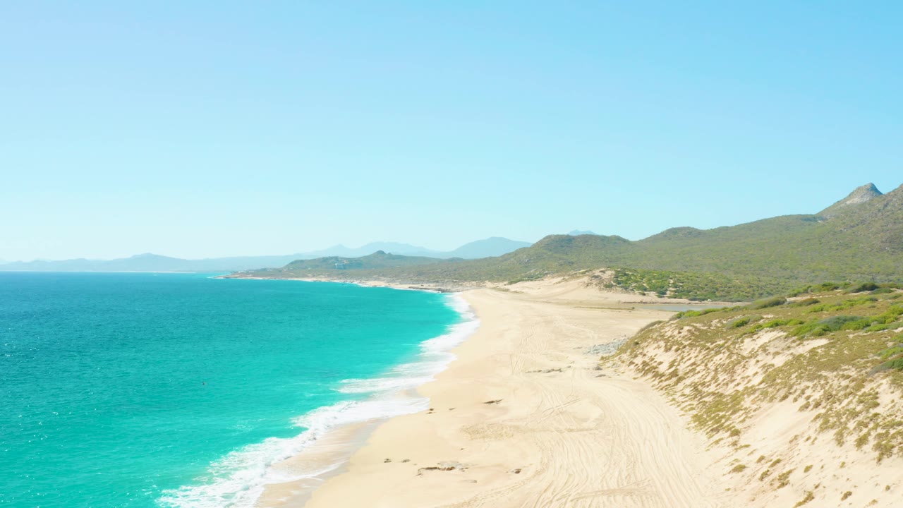East Cape BCS Mexico coastline from above, with the turquoise ocean meeting a pale beach, scrub-covered dunes on the right, green hills beyond, and small people appearing as dark marks on the sand.