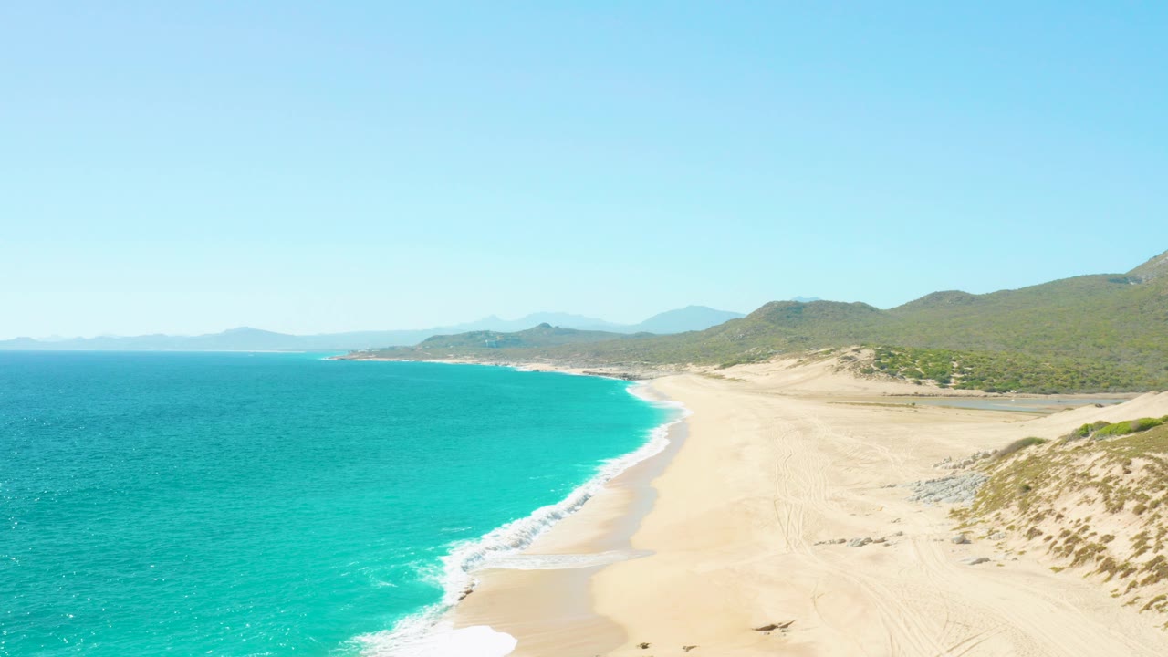 Aerial view of turquoise ocean beside a wide sandy beach and green hills.