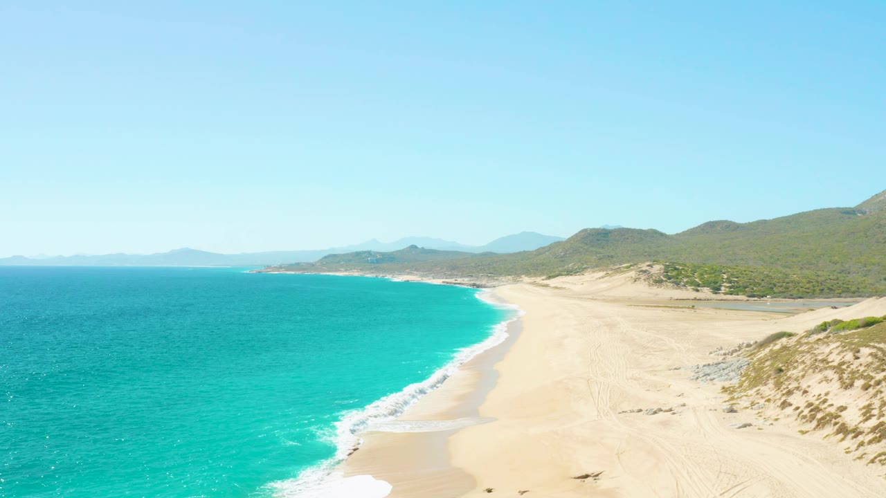 Aerial frame from East Cape BCS Mexico with the surf line slightly farther along the beach, open turquoise water to the left, dune-backed sand to the right, and no visible people.