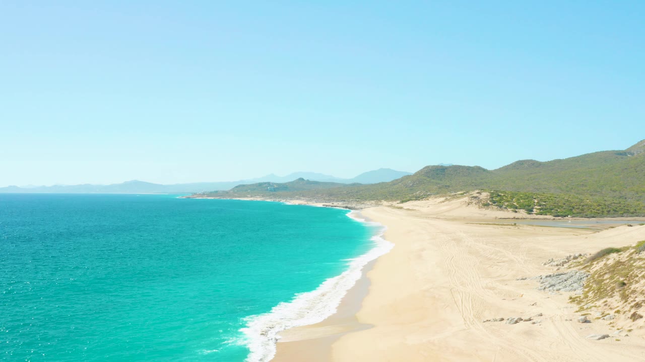Aerial frame from East Cape BCS Mexico where the ocean occupies more of the left foreground, the surf edge arcs toward the lower center, sand stretches to the right, and no visible people appear.