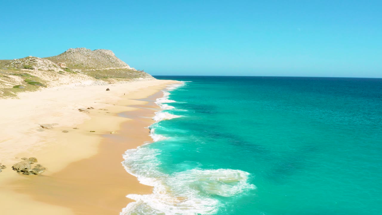Aerial view of sandy beach, turquoise surf, and low coastal hillside.