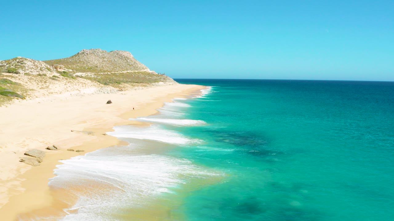 East Cape BCS Mexico beach from a high vantage, with turquoise water on the right, white surf spreading over pale sand, low hills in back, and tiny distant beach figures visible.