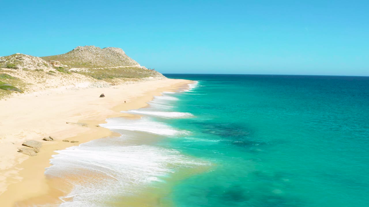 White waves wash farther up the sand beside clear turquoise water.